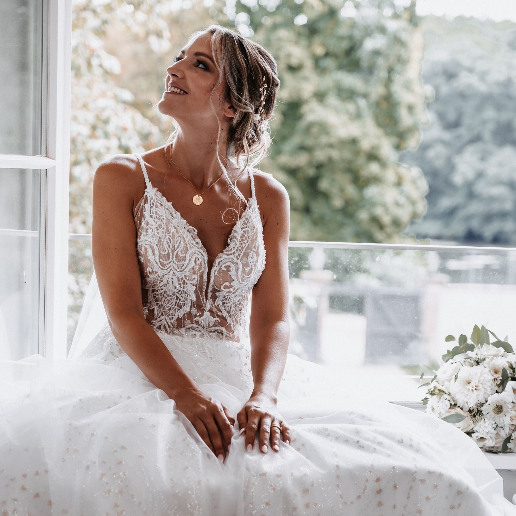 Beautiful bride in a lace wedding gown sits by a window, exuding elegance and joy.